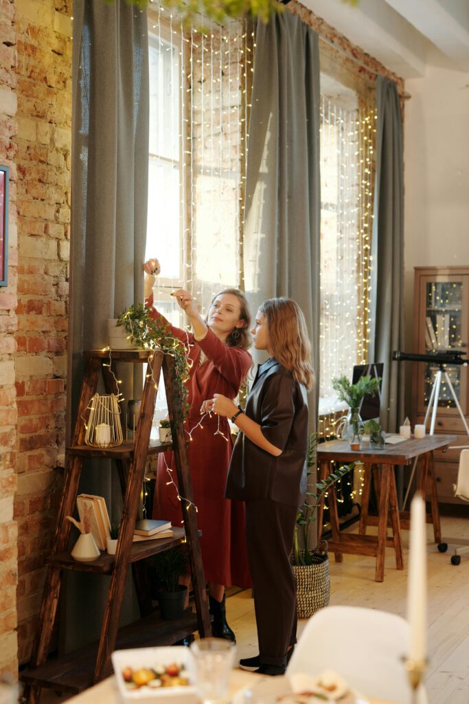 A mother and teenage daughter decorating a cozy room with Christmas lights and garlands.